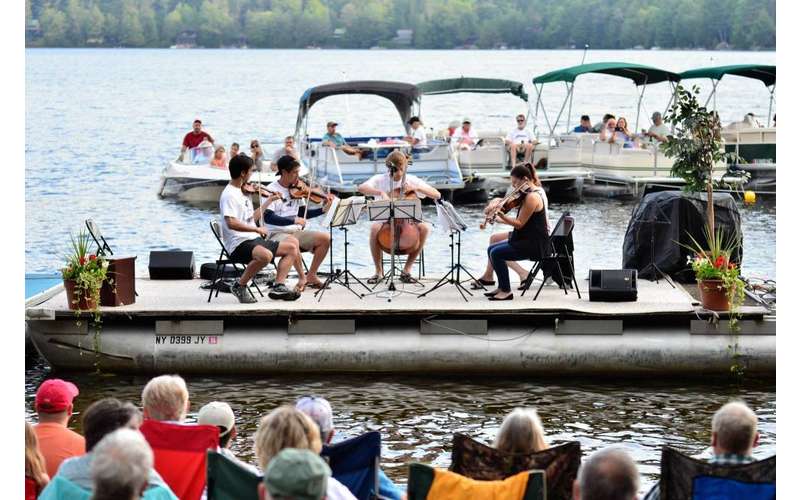 a four-piece group with string instruments perform on a boat in an open setting with other people on boats in the water watching them