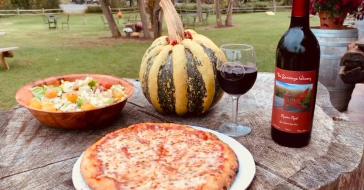 pizza, salad, wine bottle, wine glass, and squash on a wooden table outdoors