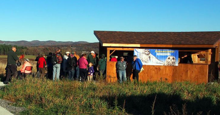 people gathered by a small hut with a Washington County Grasslands IBA poster on it