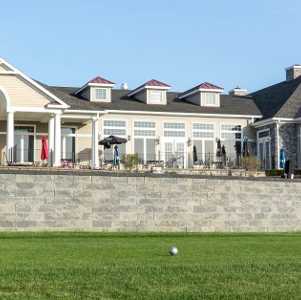 A large golf clubhouse with multiple windows, a stone retaining wall, and a well-manicured lawn under a clear blue sky.