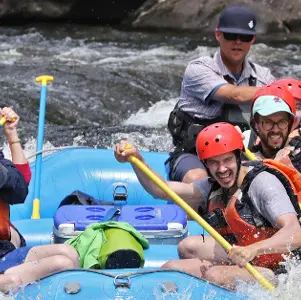 A group of people wearing helmets and life jackets paddle a blue inflatable raft through whitewater rapids.