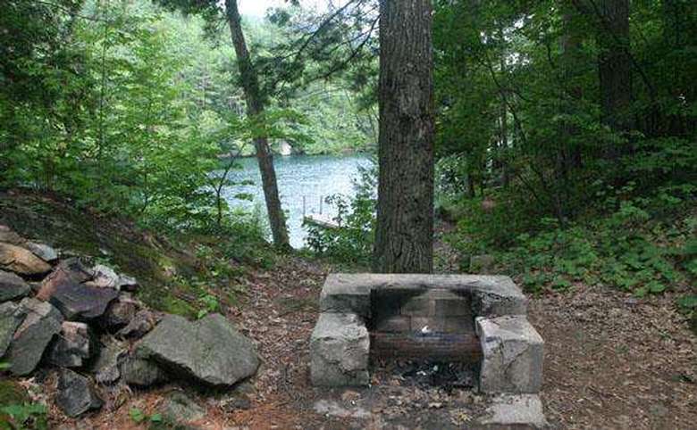a stone fire pit and grill area at a campsite in the woods with the lake in the background