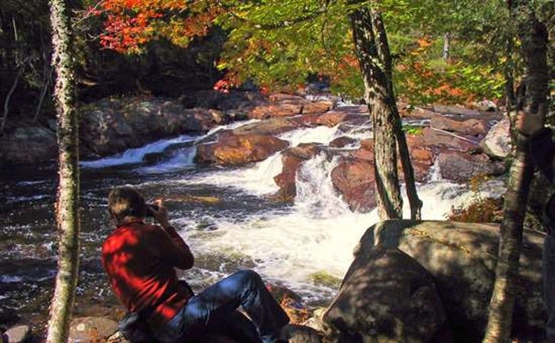 man in a red shirt taking a picture of water rushing over rocks