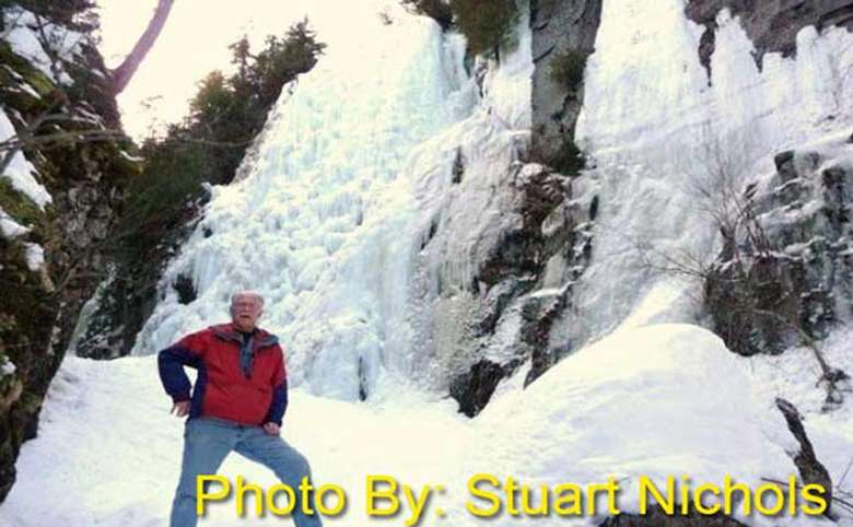 frozen waterfall in the woods with a man standing in front of it, photo credit to stuart nichols