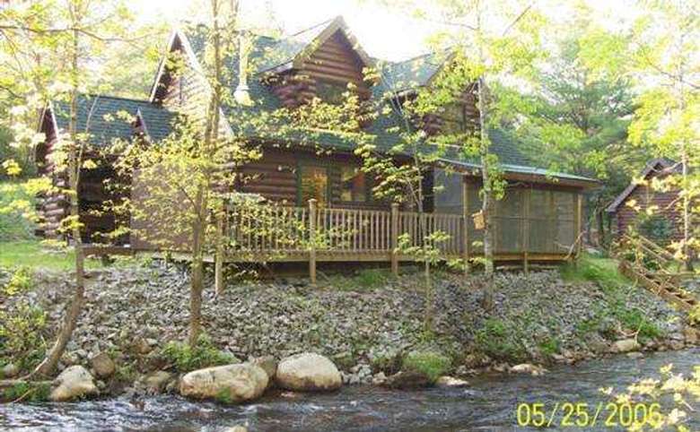 two-story log house in the woods next to a creek