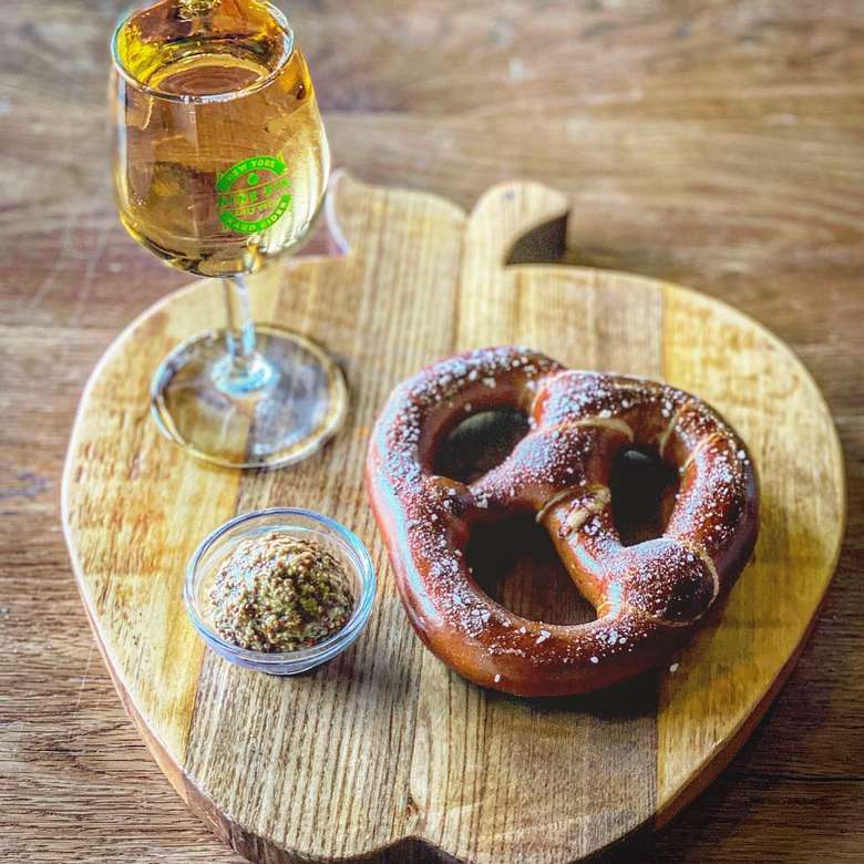 An apple shaped board with a soft pretzel, mustard, and a glass of cider laying on top