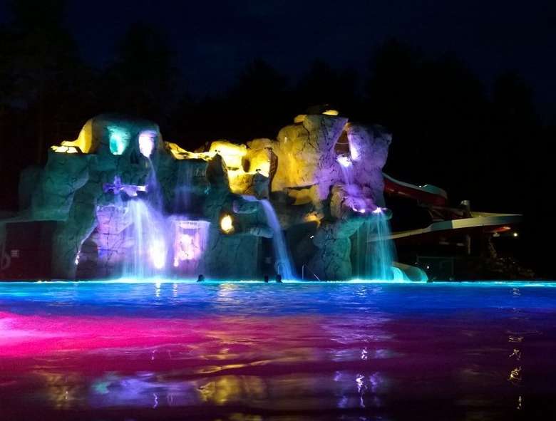 an outdoor swimming pool area with stone attractions in the back, waterfalls, and colored lights at night