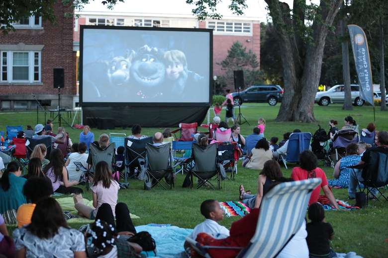 lawn full of people in chairs and on blankets watching a movie projected onto a large screen