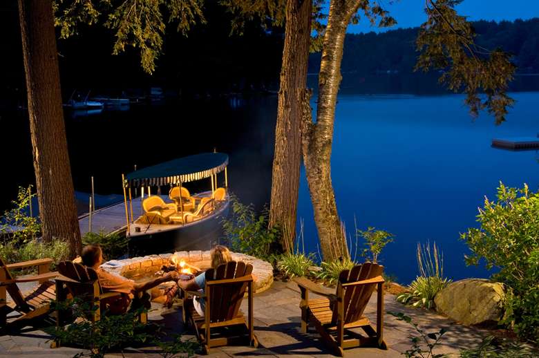 adirondack chairs in front of a firepit overlooking a docked boat and friends lake at dusk