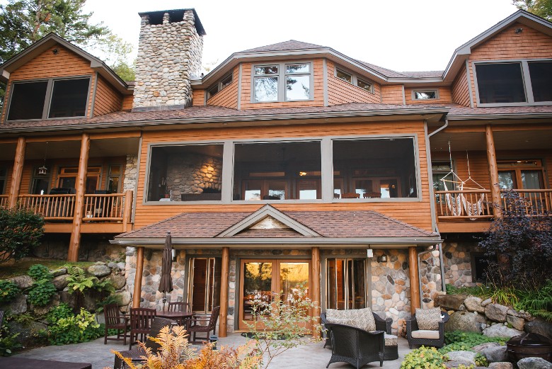 the large rear of the lodge that faces the lake and the patio below with hot tub and gardens cascading down to the lake