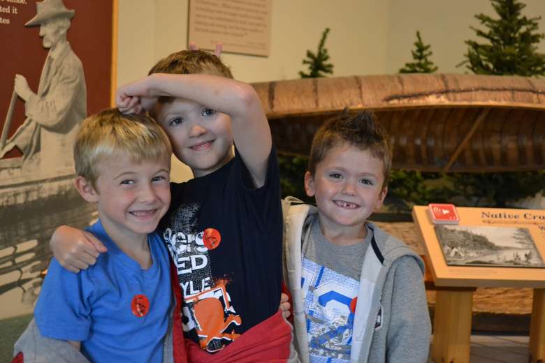 three young boys hugging each other and smiling at the camera. There is a boat display in the background.