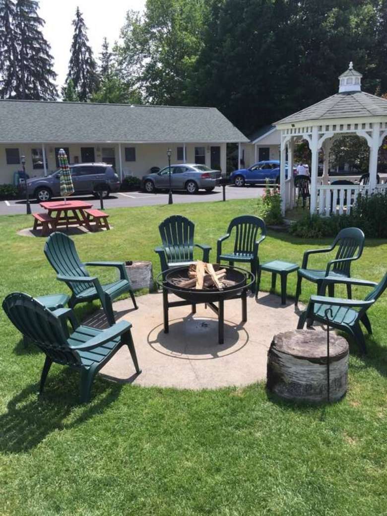 A raised fire pit surrounded by green chairs and small tables. There is a white gazebo, picnic table and lodging units in the background.
