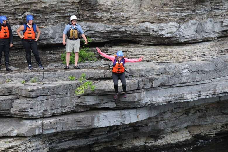 woman in a life jacket and helmet jumping off a cliff while others look on