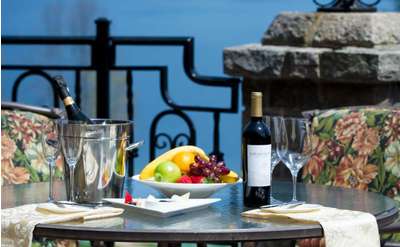 A round outdoor dining table, set on the porch of the Inn at Erlowest. You can see the water through the wrough-iron railing. The table is set with bottles of wine, glasses and a bowl of fruit.
