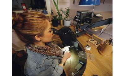 Laura sits at her bench with a microscope with her hair in a bun. behind her are several plants, on the solid wooden bench she works at. she is holding a ring in her hand and has a graver in the other demonstrating stone setting techniques