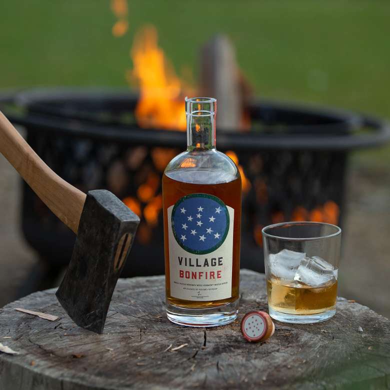 bottle of Village Bonfire by a whiskey glass and axe with a campfire in the back