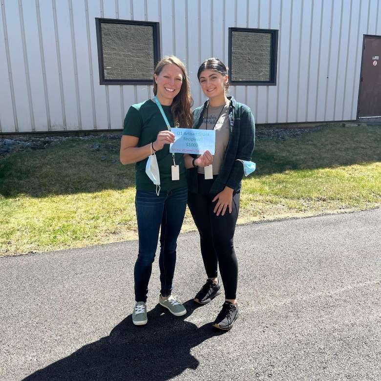 two women standing together outside building and holding up a small grant check