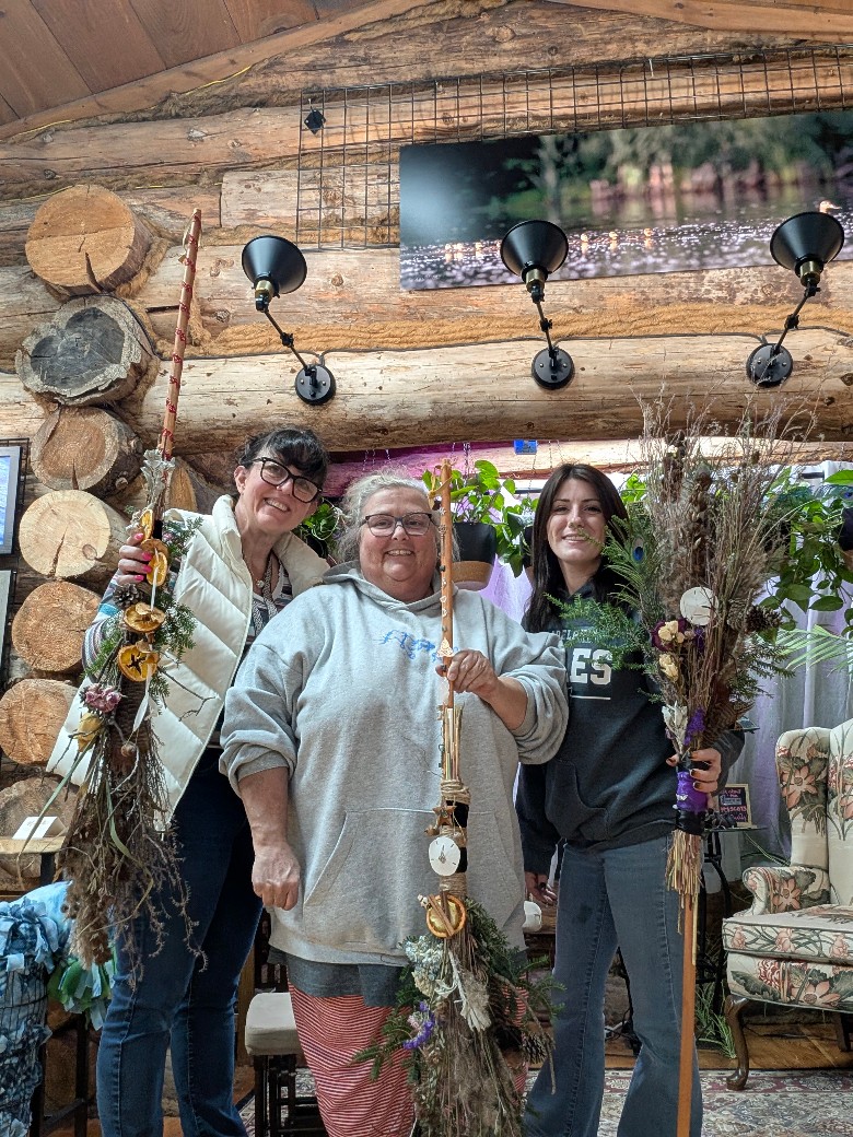 three women holding handmade brooms in a log cabin