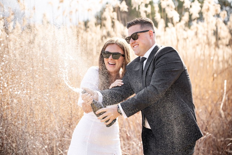 bride and groom shaking champagne bottle outdoors near field of grass