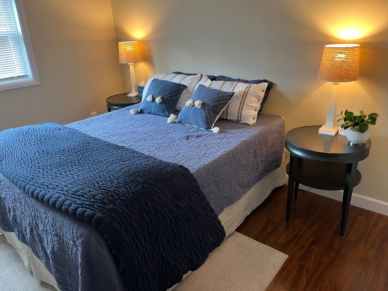 a house bedroom with a bed, blue covers, four pillows, and two nightstands with lamps turned on