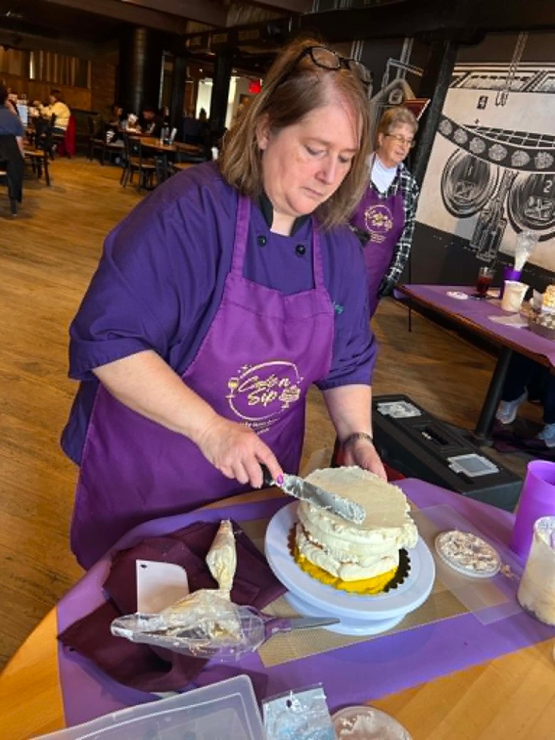 a woman wearing a purple apron putting buttercream on a cake