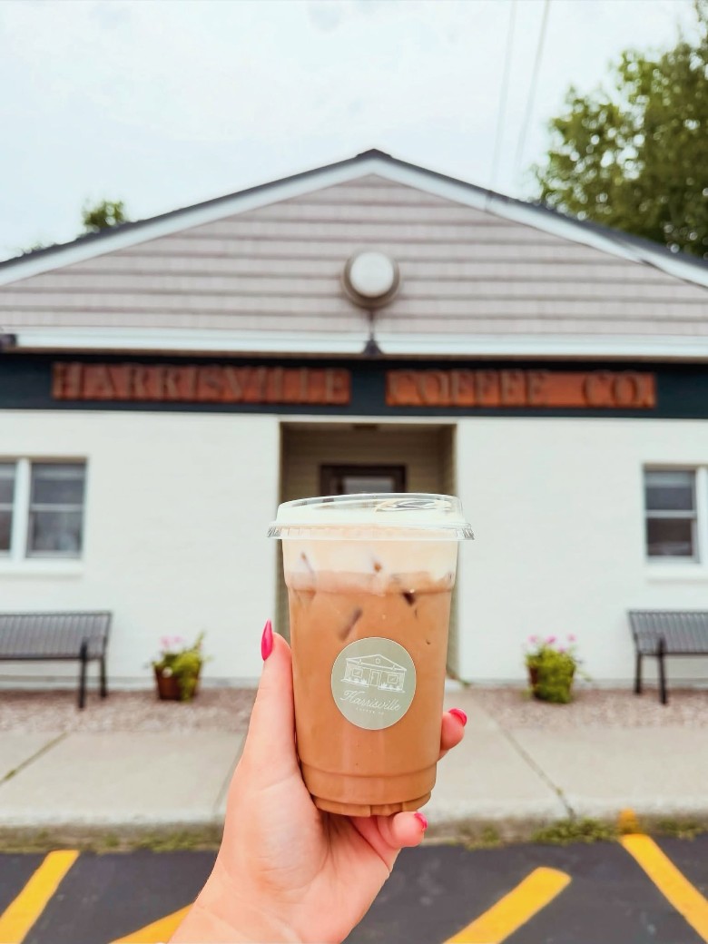 The exterior of Harrisville Coffee Co., housed in a white building. a person holding up an iced coffee in front of a coffee shop