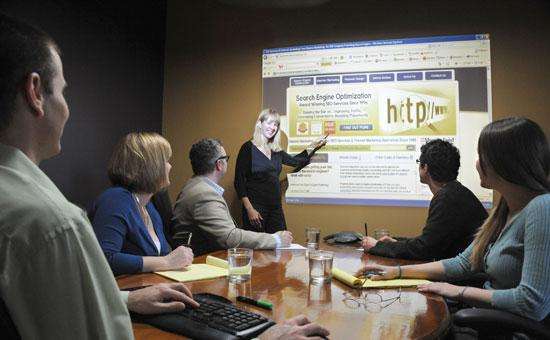 a woman speaking and pointing at a projector screen in a conference room