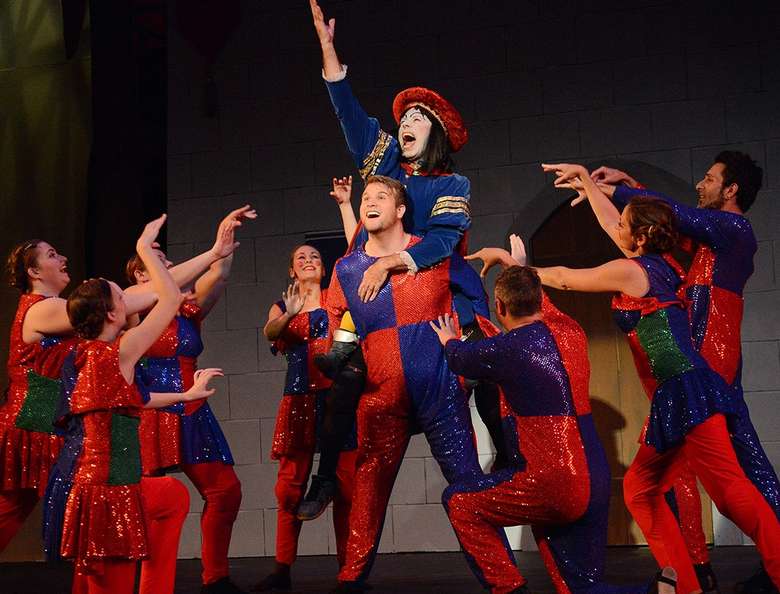 actor playing lord farquad in shrek singing with other actors in red and blue costumes