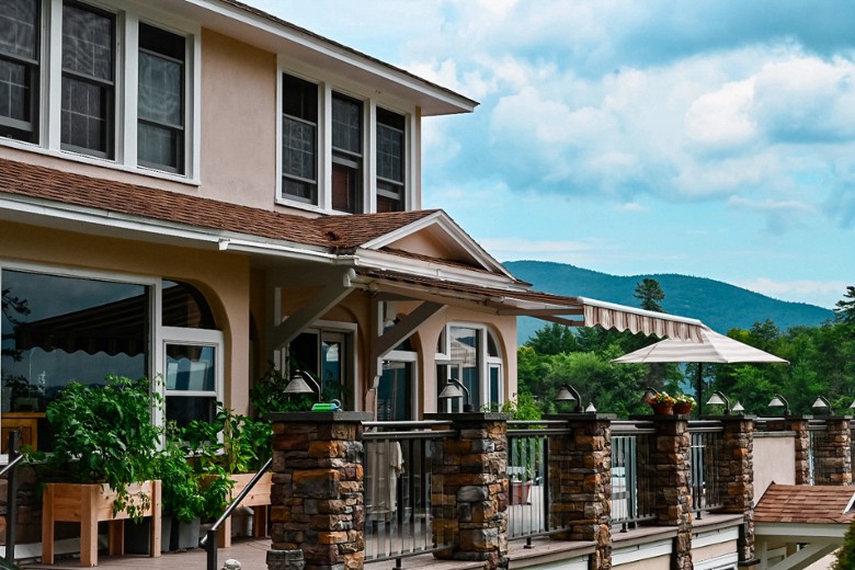 A beige two-story house features a spacious deck with stone pillars, potted plants, and a scenic view of forested mountains under a partly cloudy sky.