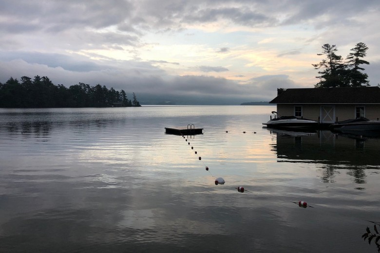 A still lake at dawn reflects soft clouds and distant trees, with a floating dock and a boathouse along the shore.
