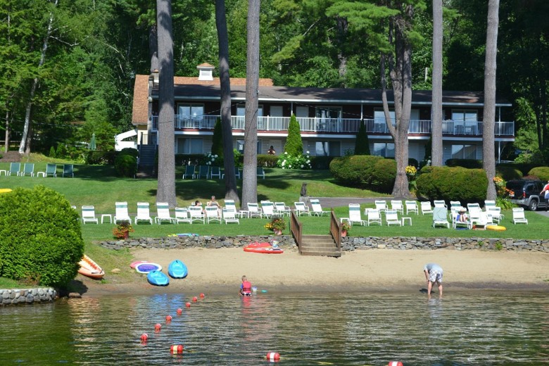 A lakeside lodge sits behind a manicured lawn with lounge chairs and kayaks, while people enjoy the sandy beach and shallow water in the foreground.
