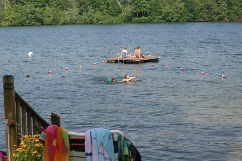 Children swim in a designated lake area while two teens relax on a floating dock, with colorful towels draped over a nearby railing.