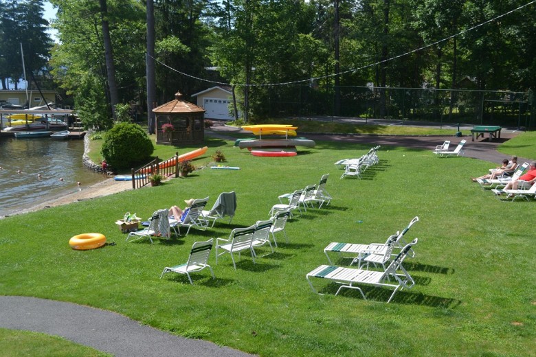 Lounge chairs are arranged on a sunny lakeside lawn near a small beach, gazebo, kayaks, and a fenced recreation area.