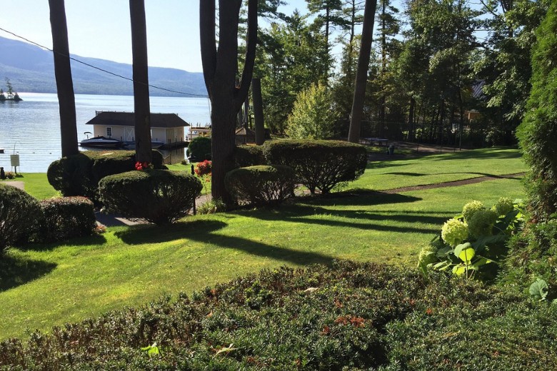 A well-kept lawn with trimmed bushes and blooming flowers overlooks a peaceful lake and boathouse, framed by tall trees and distant mountains.