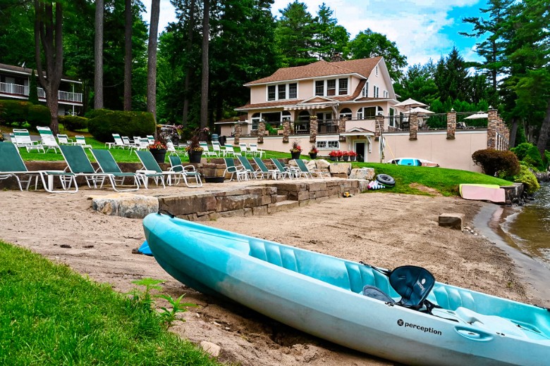 A light blue kayak rests on the sandy beach in front of a lakeside house surrounded by lounge chairs, greenery, and tall trees.