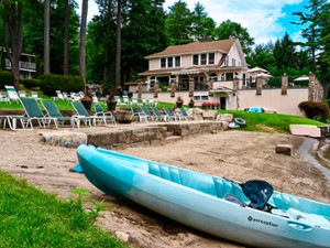 A light blue kayak rests on the sandy beach in front of a lakeside house surrounded by lounge chairs, greenery, and tall trees.