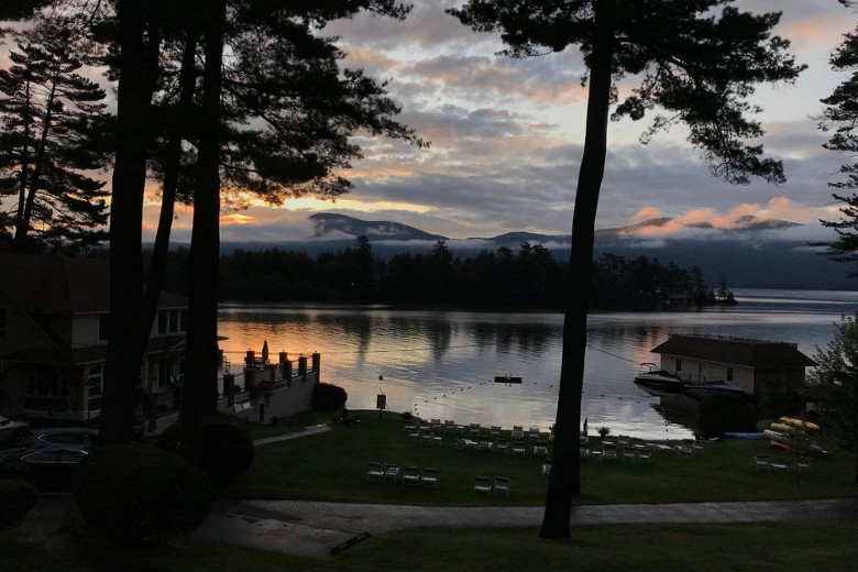 A serene lakeside view at sunrise shows silhouetted trees, lounge chairs, and a boathouse reflecting on calm water with misty mountains in the distance.