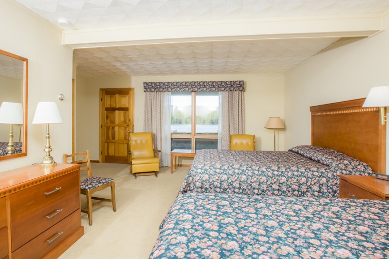 A cozy hotel room features two floral-patterned double beds, wooden furniture, and a window with lake and mountain views.