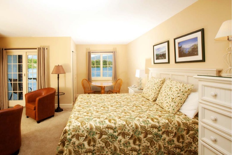 A bright and inviting guest room features a king-sized bed, cozy seating, and large windows with views of a scenic lake and forested hills.