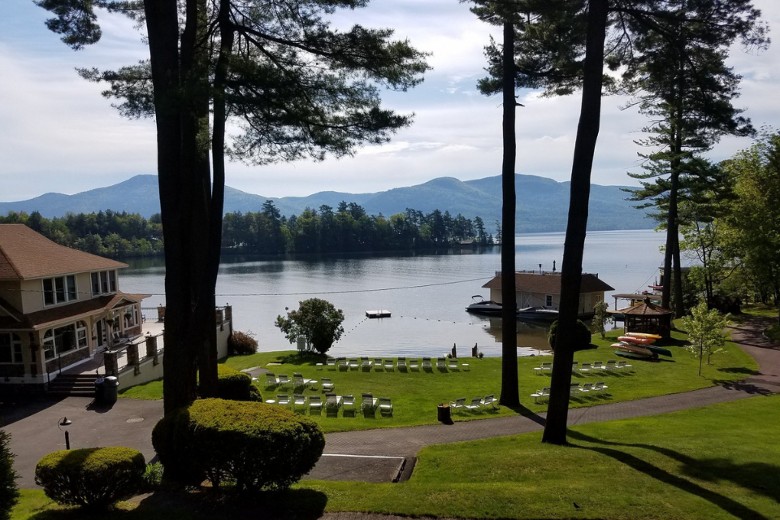 A lakeside property features a large house, lounge chairs on a green lawn, and a boathouse on calm water with mountains in the background.