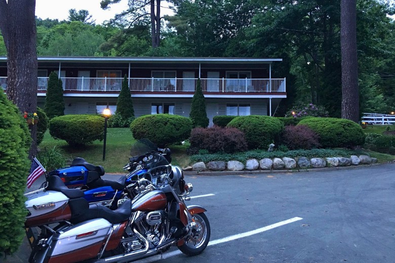 Two touring motorcycles are parked in front of a two-story motel surrounded by manicured shrubs and tall trees at dusk.