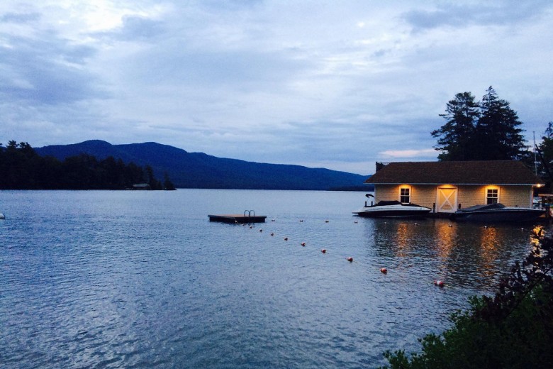A softly lit boathouse reflects on the lake at dusk, with a floating dock and dark mountain silhouettes in the background.