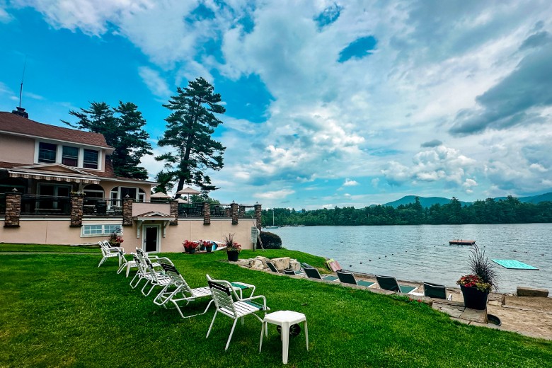 Lounge chairs line a grassy hill beside a lake, with a large house overlooking the water and mountains in the distance under a partly cloudy sky.