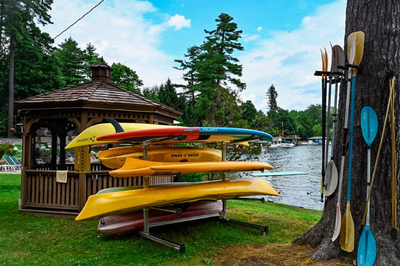 Stacked kayaks and paddles sit beside a wooden gazebo near a calm lakeshore lined with trees and boats.