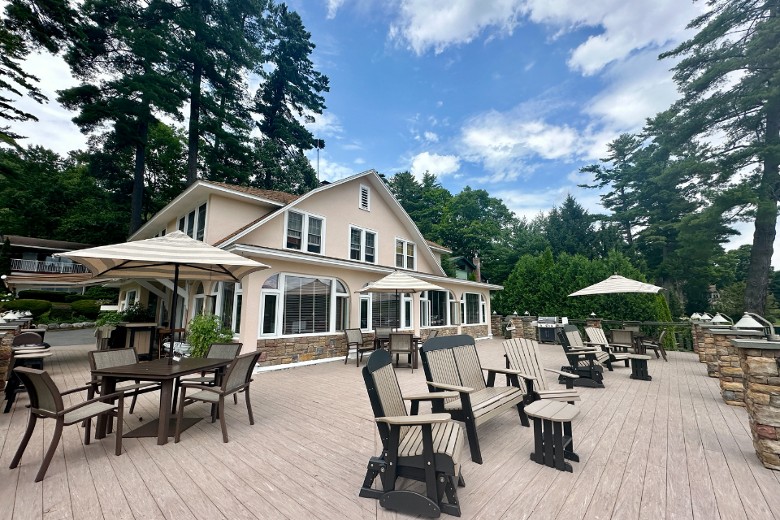 An expansive wooden deck with patio furniture and umbrellas wraps around a beige house surrounded by tall trees and blue skies.