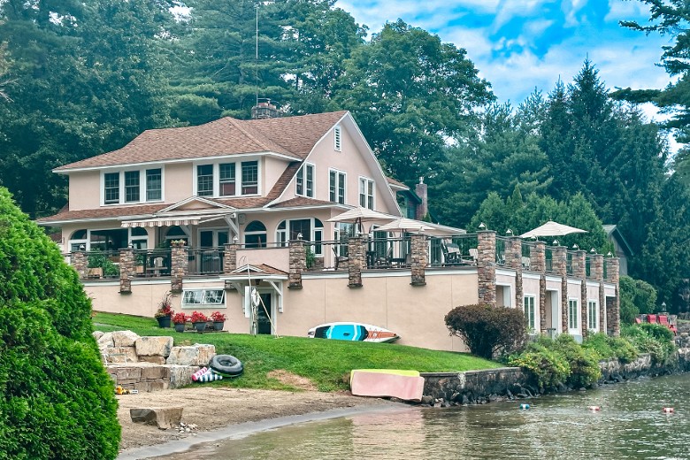 A large lakeside house with a spacious patio, stone columns, and kayaks resting on the grassy shore near calm water.