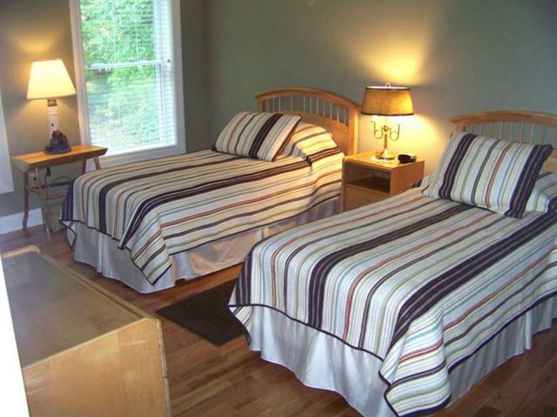 two beds with headboards, a nightstand and lamp in between, and a table with lamp by the windows to the right of the beds.