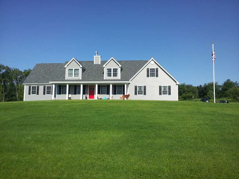 a grey two-story home on a very large grassy lot, with a front porch, red door, and two dormers in the front