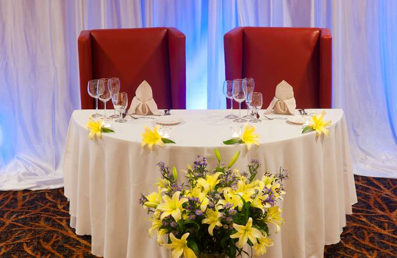 the couple's small, special table with white linens, glassware, and four yellow flowers as decorations with a larger bouquet of yellow flowers in front of the table