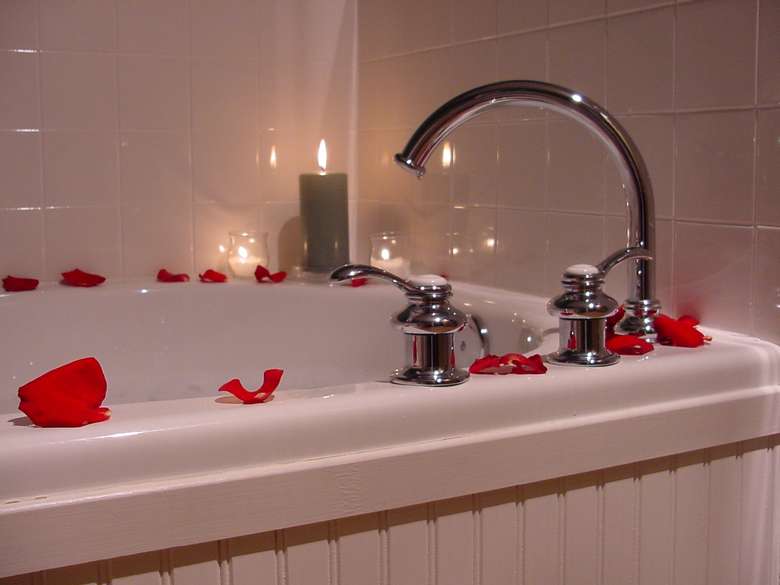large tub covered in rose petals with a lit candle on the corner of the tub reflecting light from the tub surround